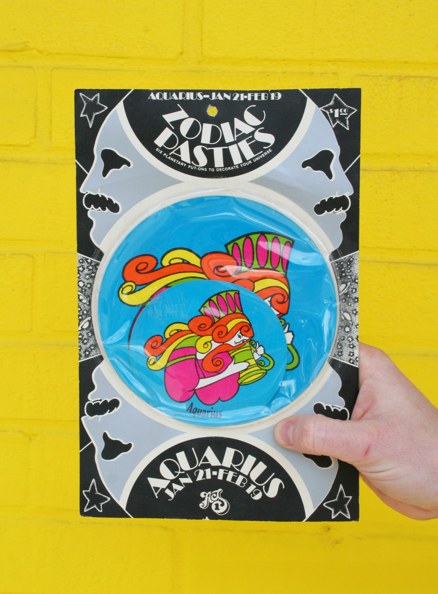A hand holding a vintage 1960s psychedelic zodiac pasty with illustrations of zodiac signs and cosmic elements against a yellow background.
