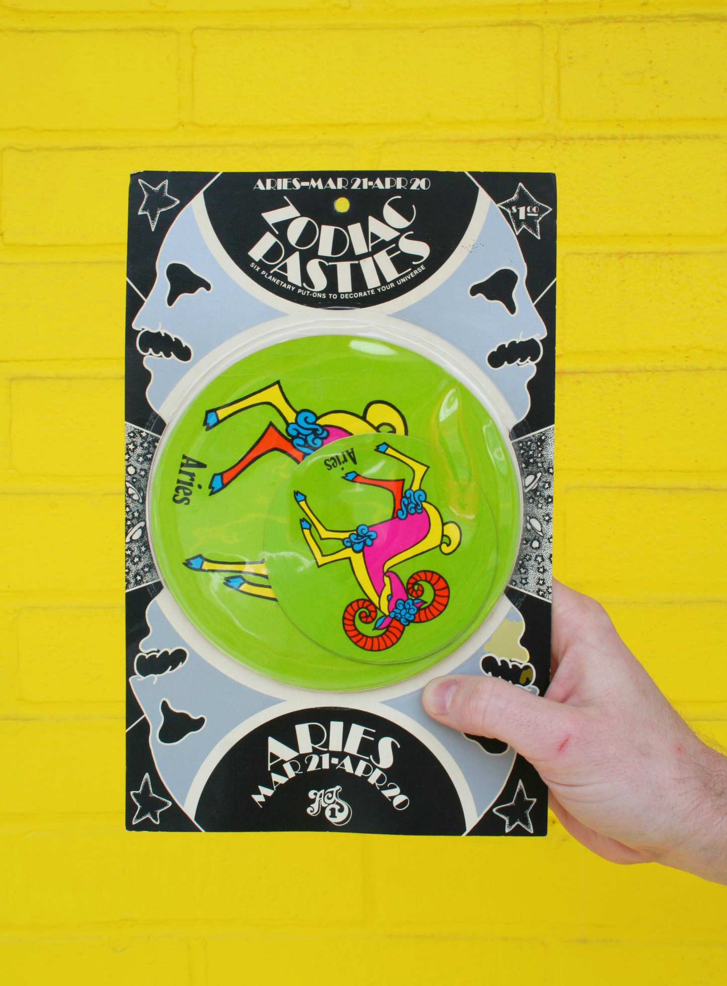 A hand holding a vintage 1960s psychedelic zodiac pasty with illustrations of zodiac signs and cosmic elements against a yellow background.
