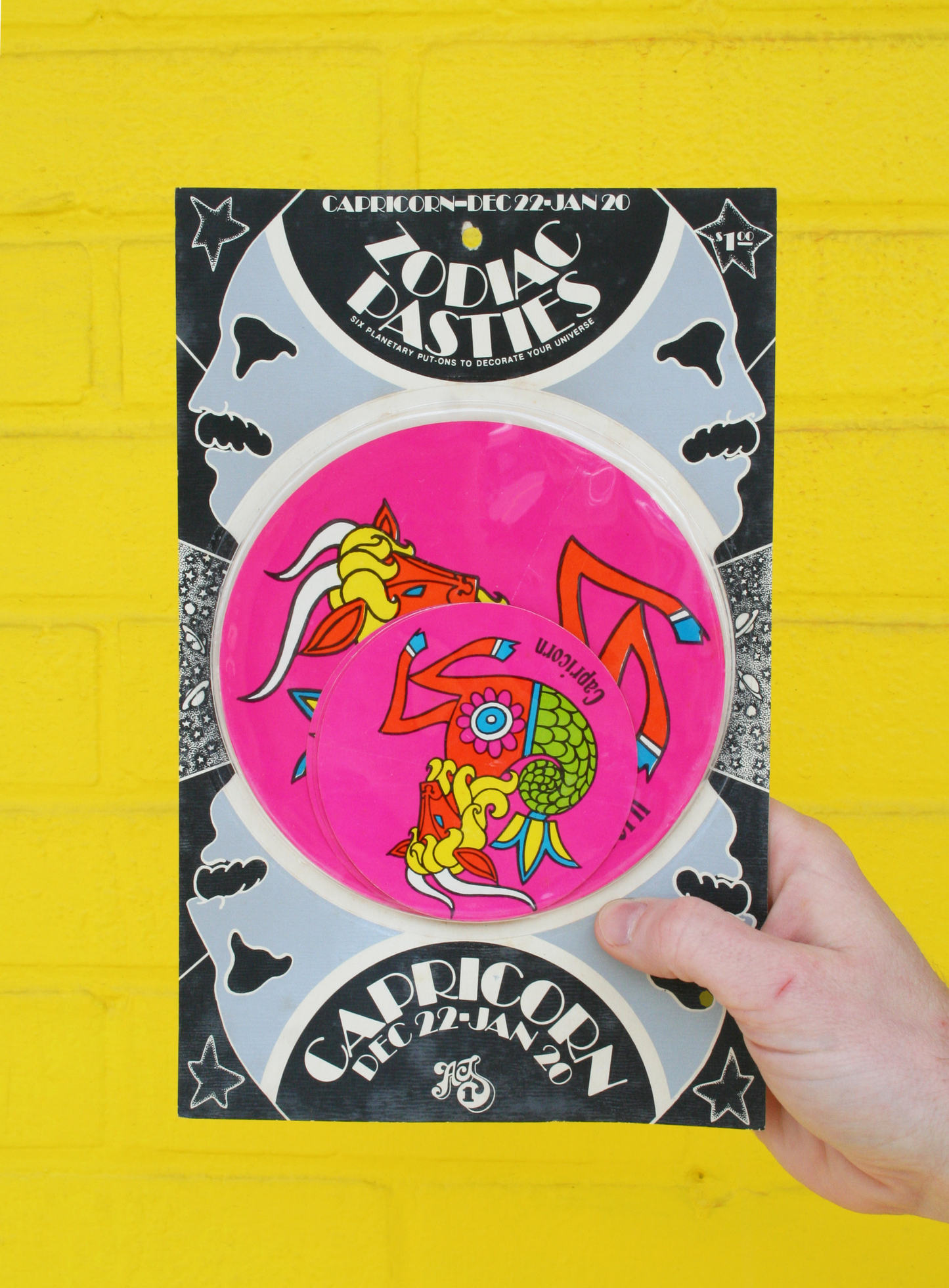 A hand holding a vintage 1960s psychedelic zodiac pasty with illustrations of zodiac signs and cosmic elements against a yellow background.