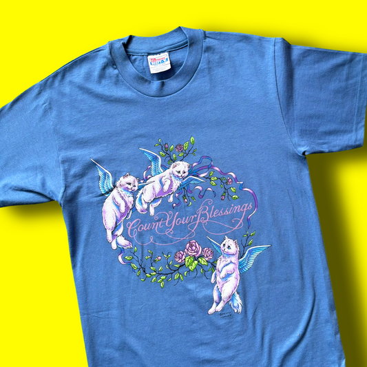 A blue unworn t-shirt with a graphic print of angel cats and the phrase 'Count Your Blessings' on a yellow background.