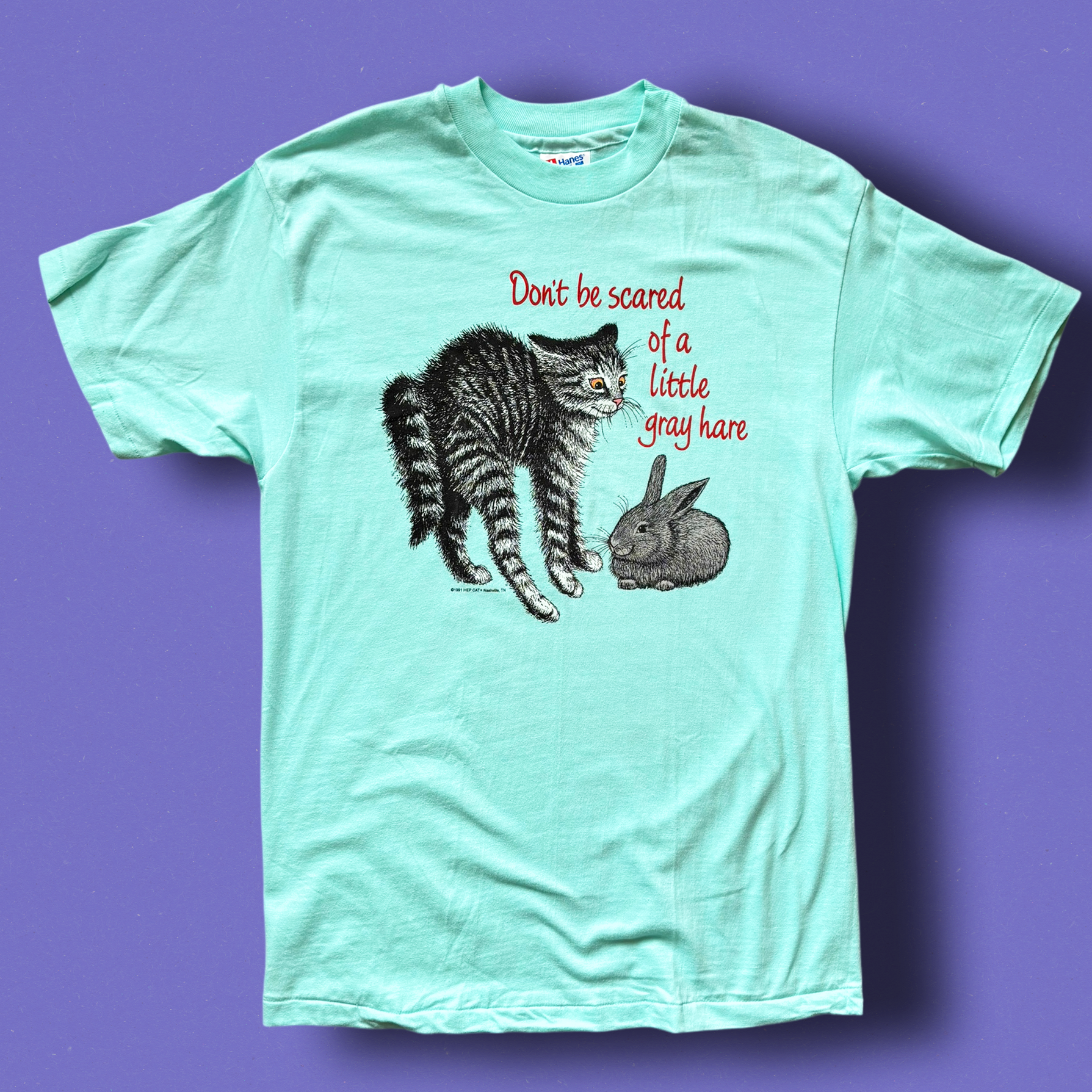 A green t-shirt with a graphic print of a cat and a rabbit, and the text 'Don't be scared of a little gray hare'. Vintage Hep Cat Tee