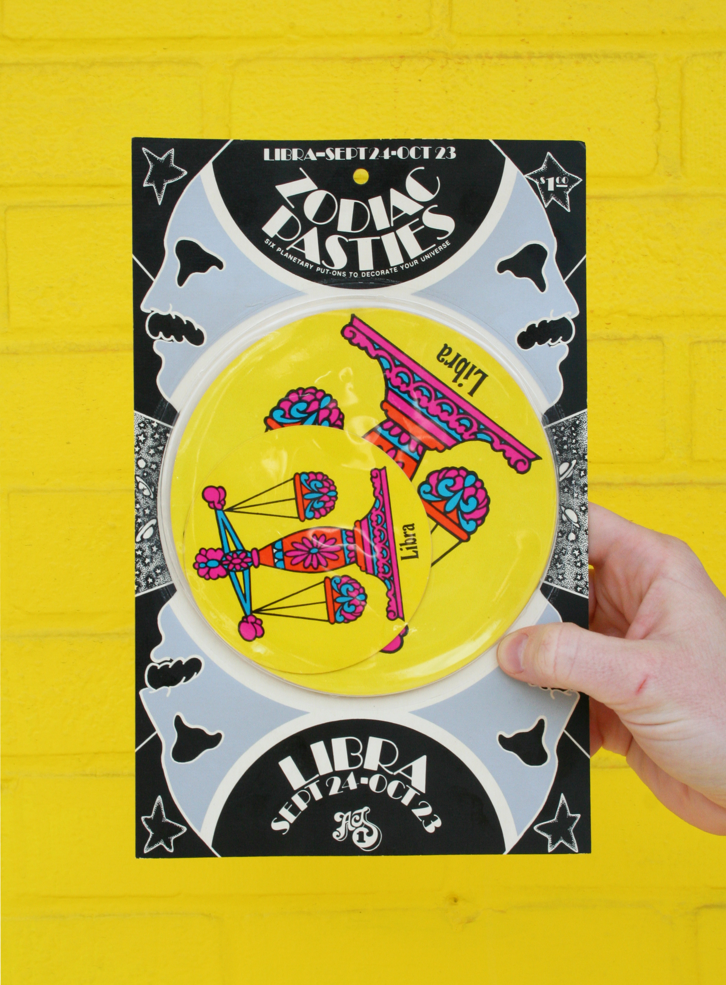 A hand holding a vintage 1960s psychedelic zodiac pasty with illustrations of zodiac signs and cosmic elements against a yellow background.