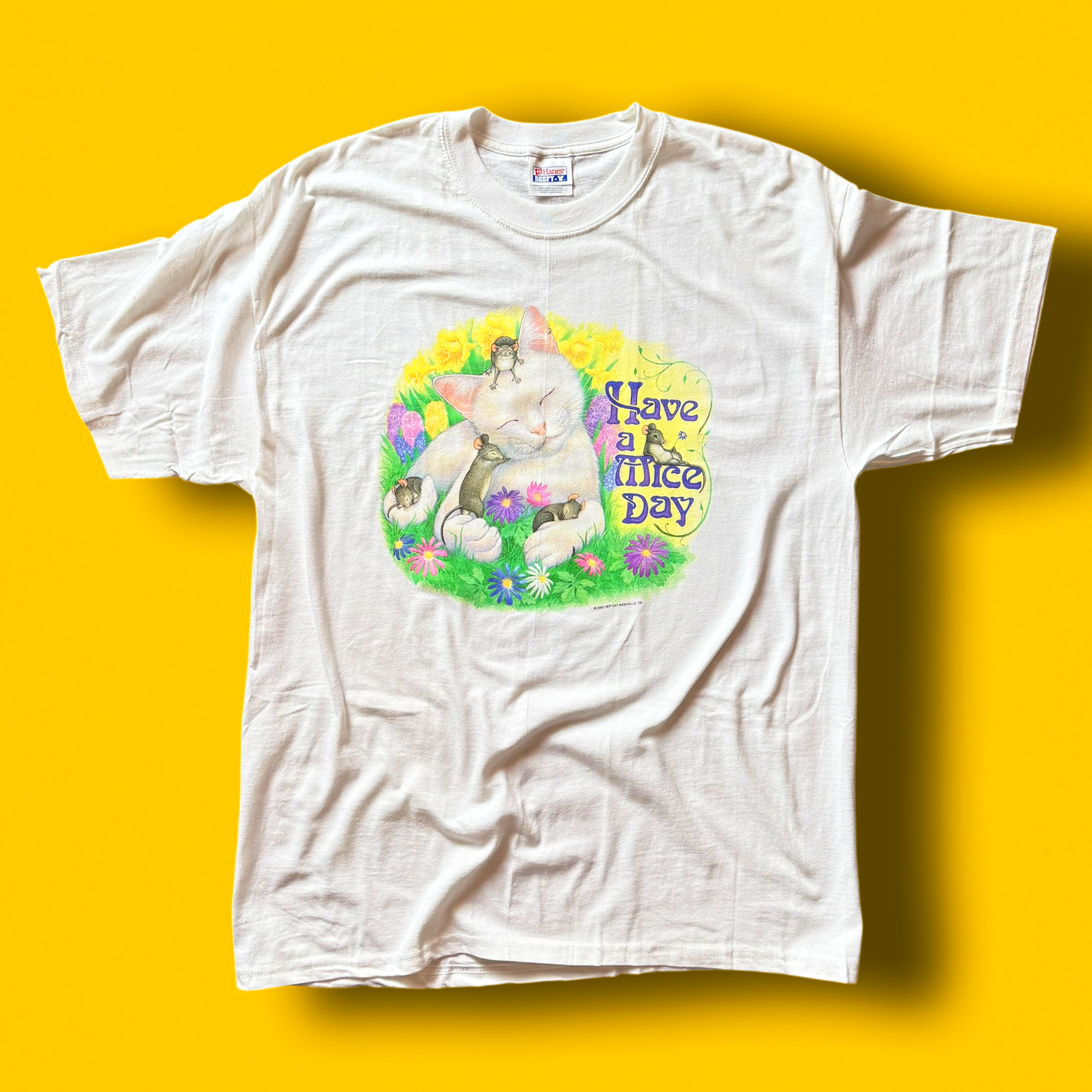 A white crew neck t-shirt with a graphic print in the center, featuring a cat, mice, and the text 'Have a Mice Day' on a yellow background.