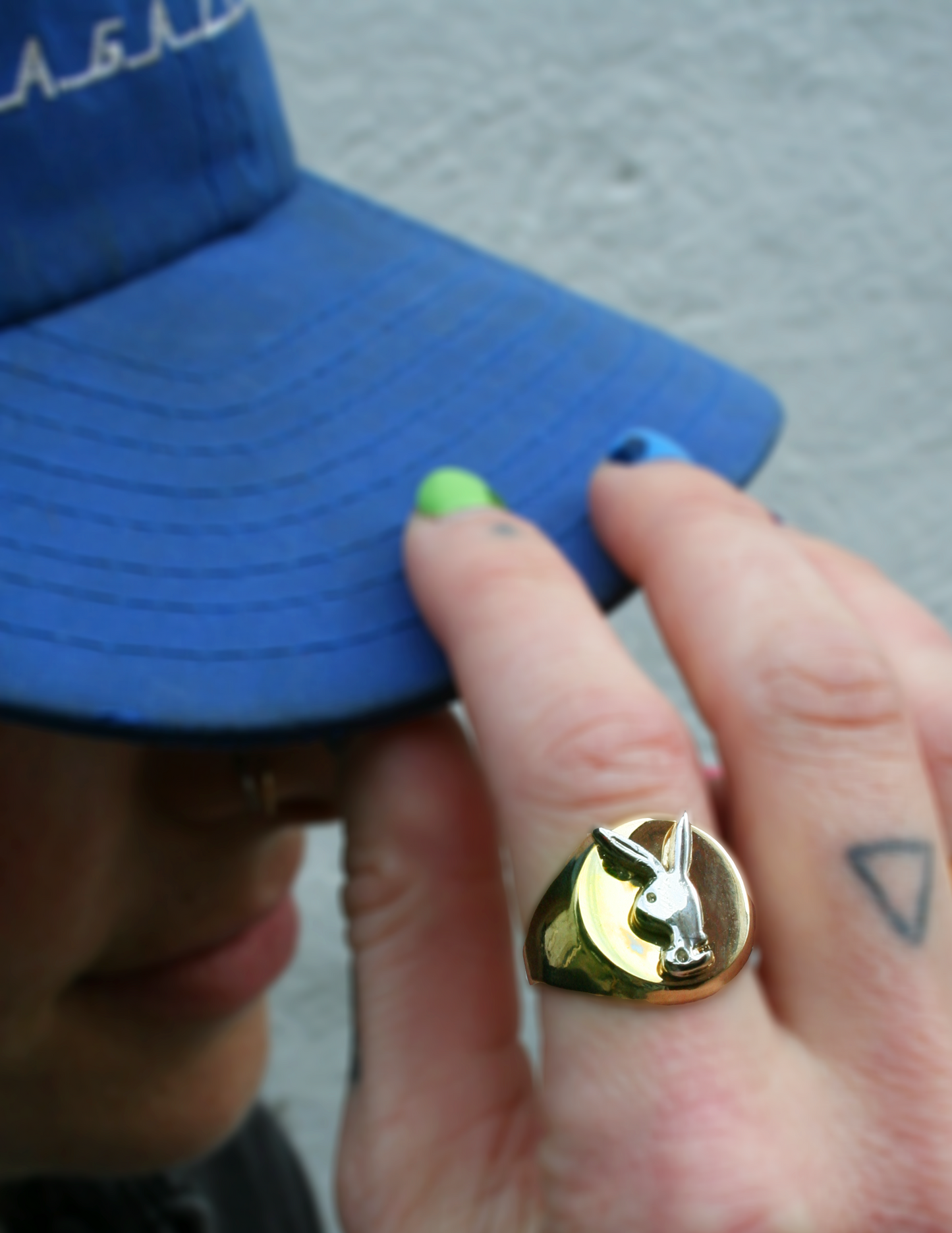 A close-up image of a person's hand wearing a gold-colored ring with a logo on it, and the same hand holding a blue hat.