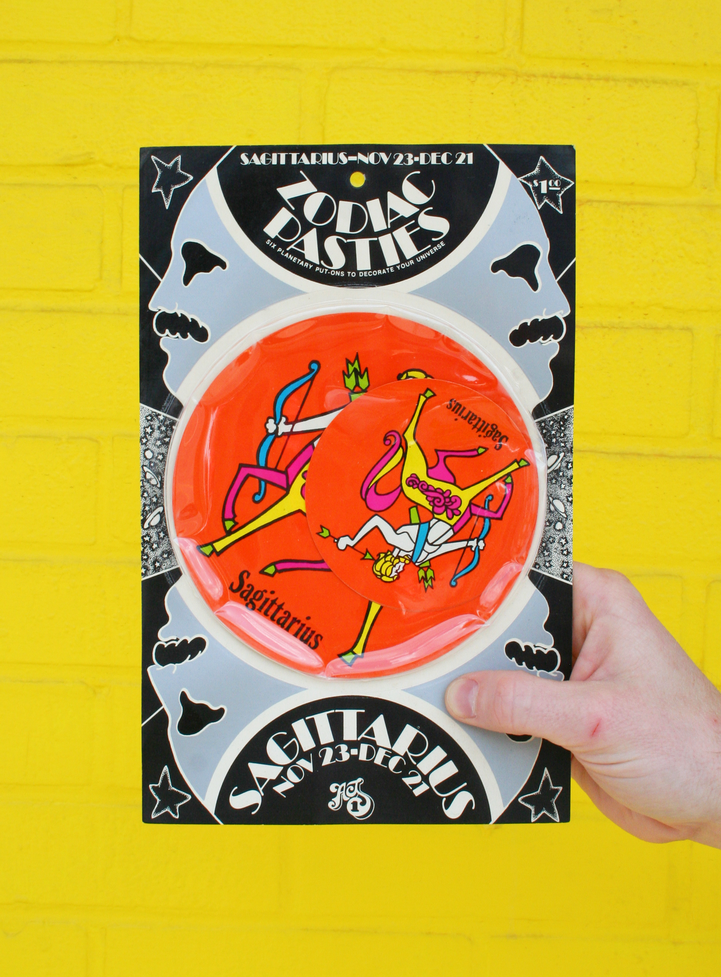 A hand holding a vintage 1960s psychedelic zodiac pasty with illustrations of zodiac signs and cosmic elements against a yellow background.