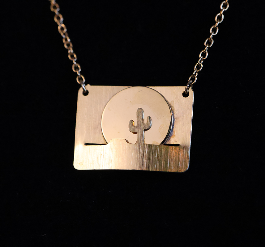 A gold-colored pendant featuring a landscape design with a sun and a cactus, attached to a chain.