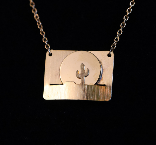 A gold-colored pendant featuring a landscape design with a sun and a cactus, attached to a chain.
