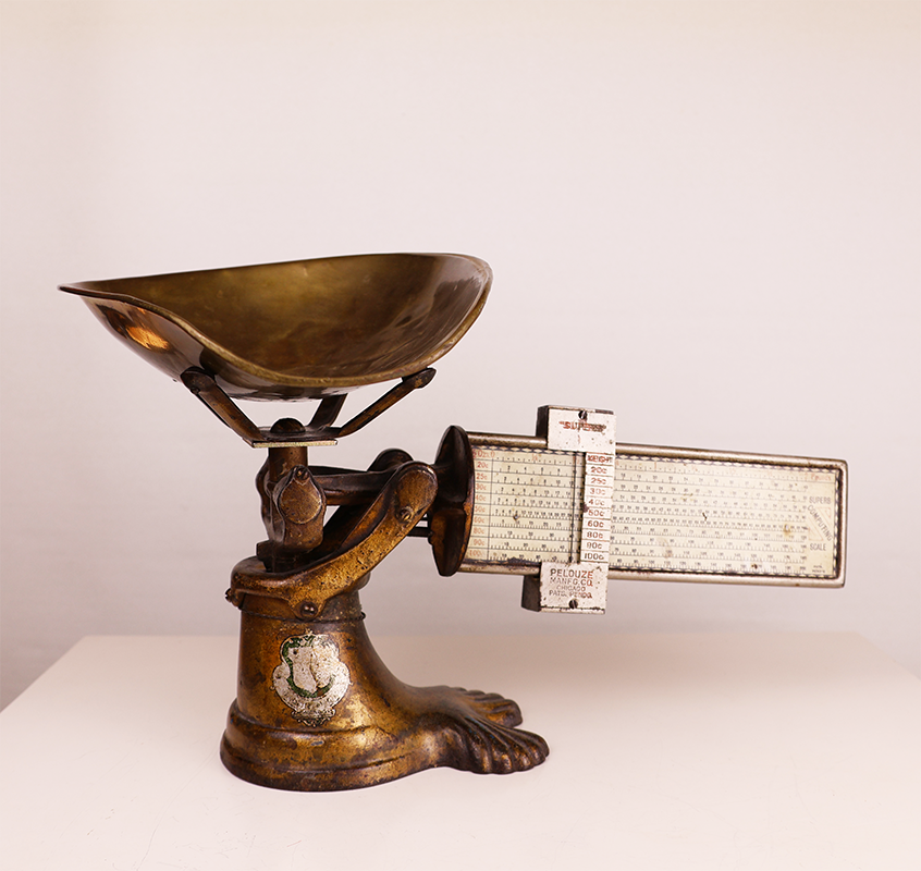 Antique brass candy scale with a measuring mechanism and a decorative base.