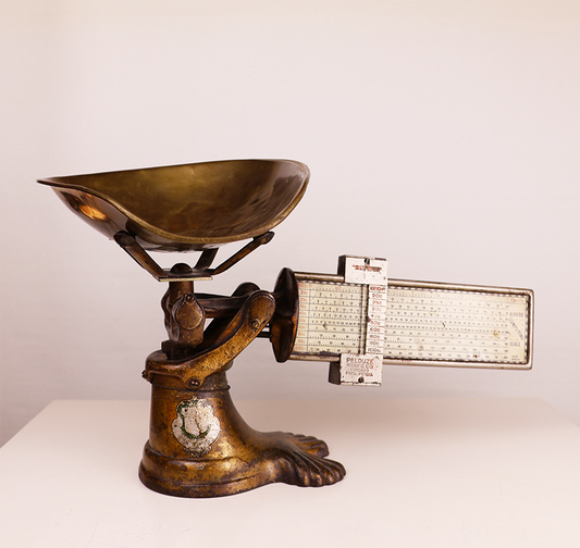 Antique brass candy scale with a measuring mechanism and a decorative base.