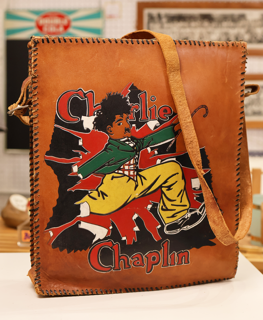 A brown leather satchel with a hand-tooled and painted Charlie Chaplin graphic in multicolor.