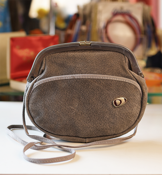 A grey suede handbag with a shoulder strap, displayed against a blurred background.