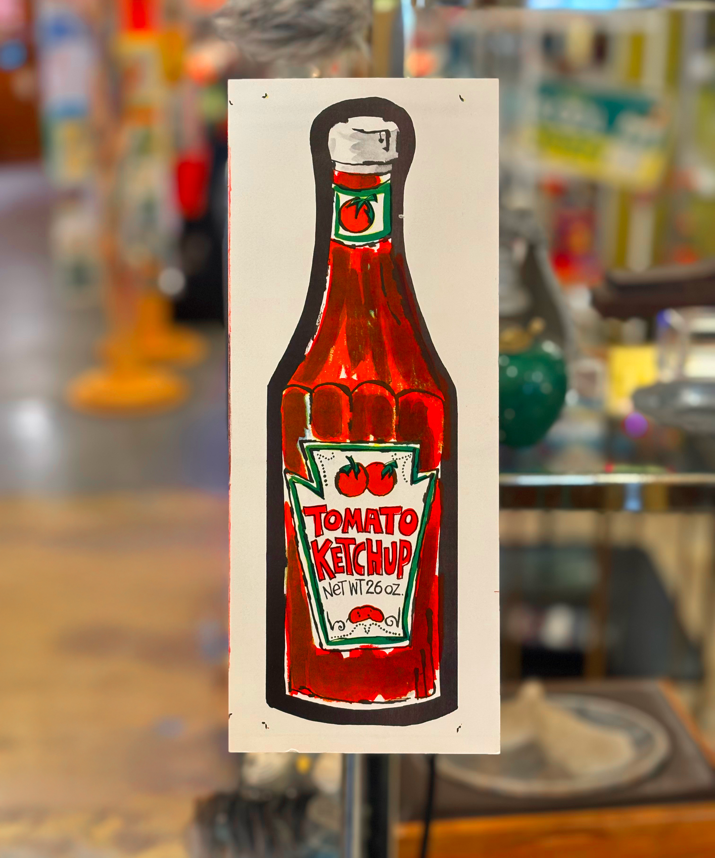 A vibrant red and green art print of a tomato ketchup bottle with a 'Tomato Ketchup' text and cherry design, displayed against a blurred background.