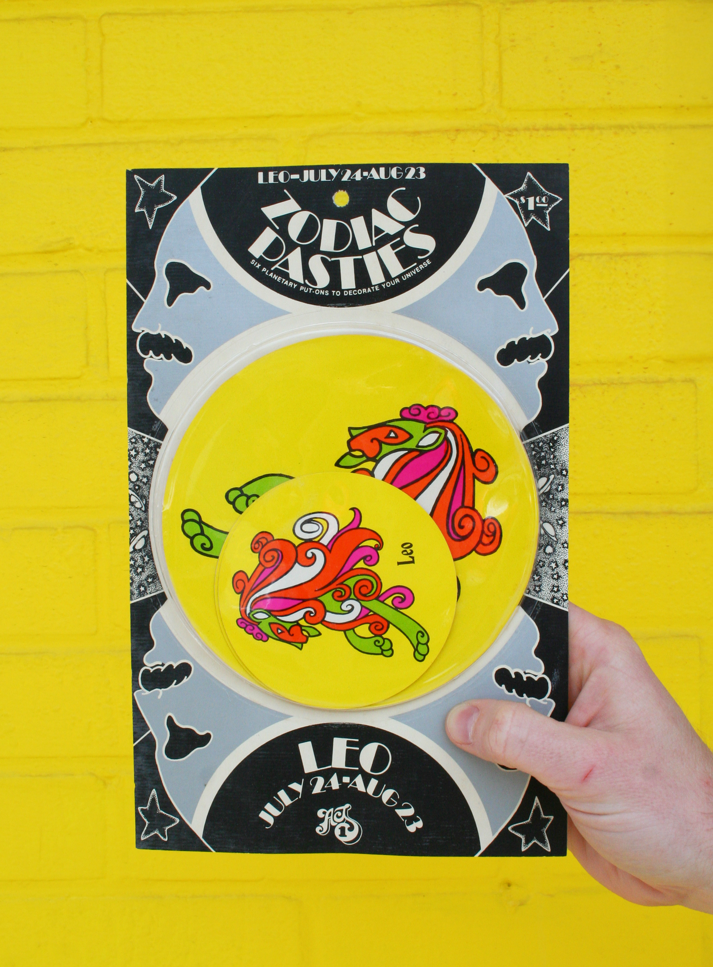 A hand holding a vintage 1960s psychedelic zodiac pasty with illustrations of zodiac signs and cosmic elements against a yellow background.