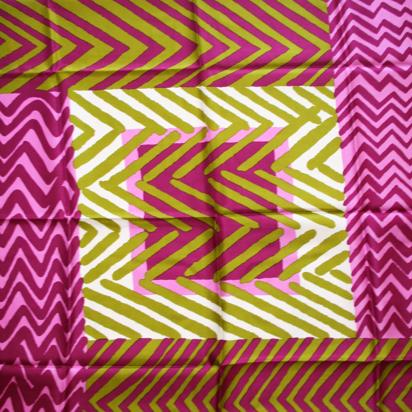 A close-up of a silk scarf with a geometric pattern in vibrant colors, featuring pink, green, and yellow stripes on a white background.