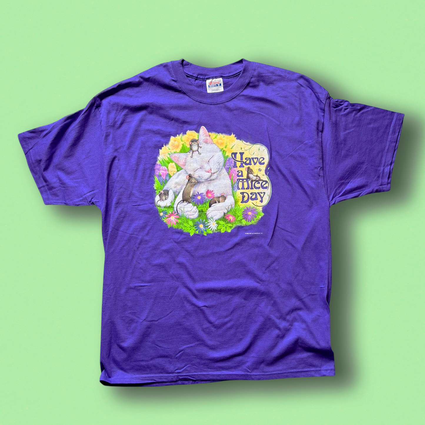 A purple crew neck t-shirt with a graphic print in the center, featuring a cat and the text 'Have a Mice Day' on a green background.