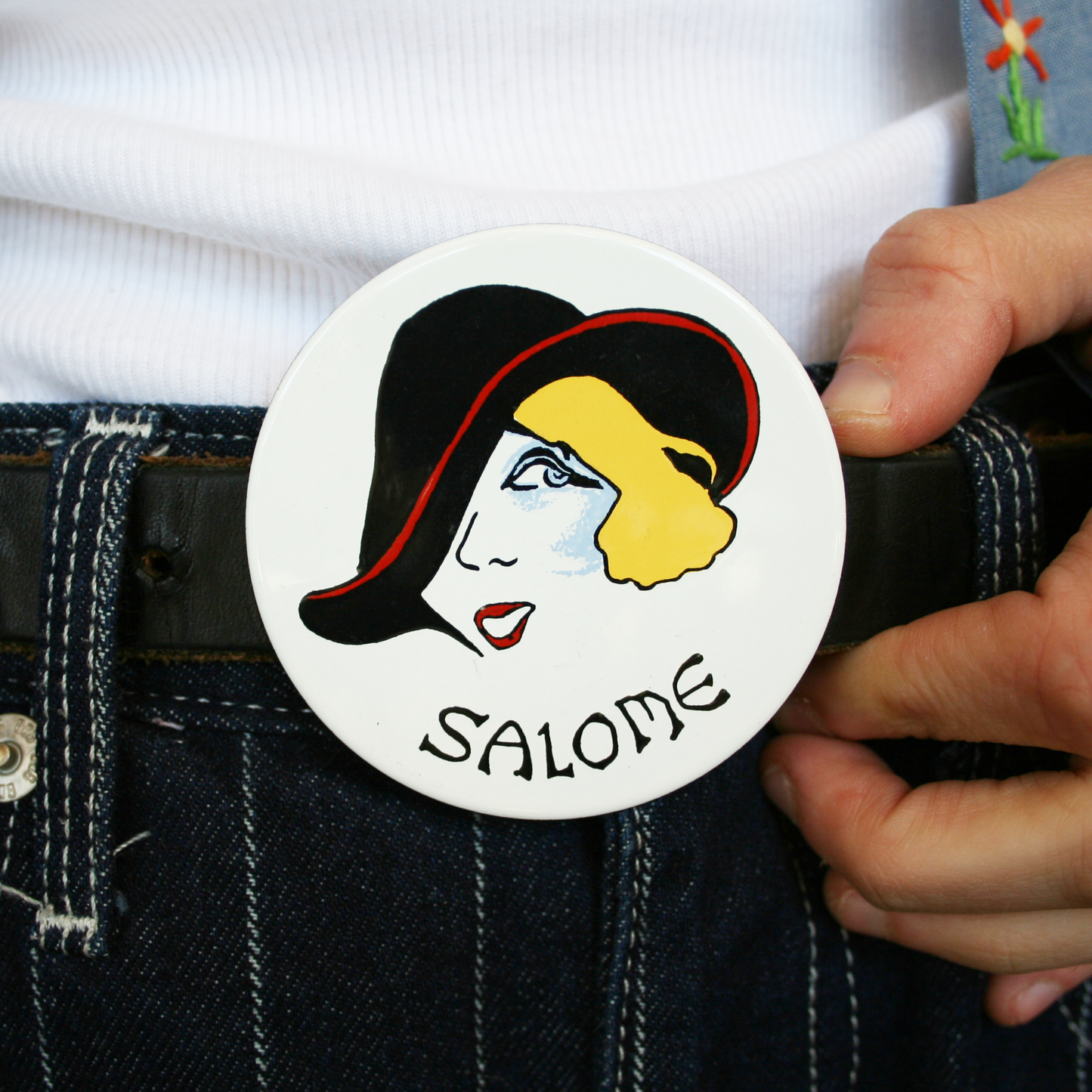 A hand holding a belt buckle with a vintage illustration of a woman named Salome wearing a black hat and white background