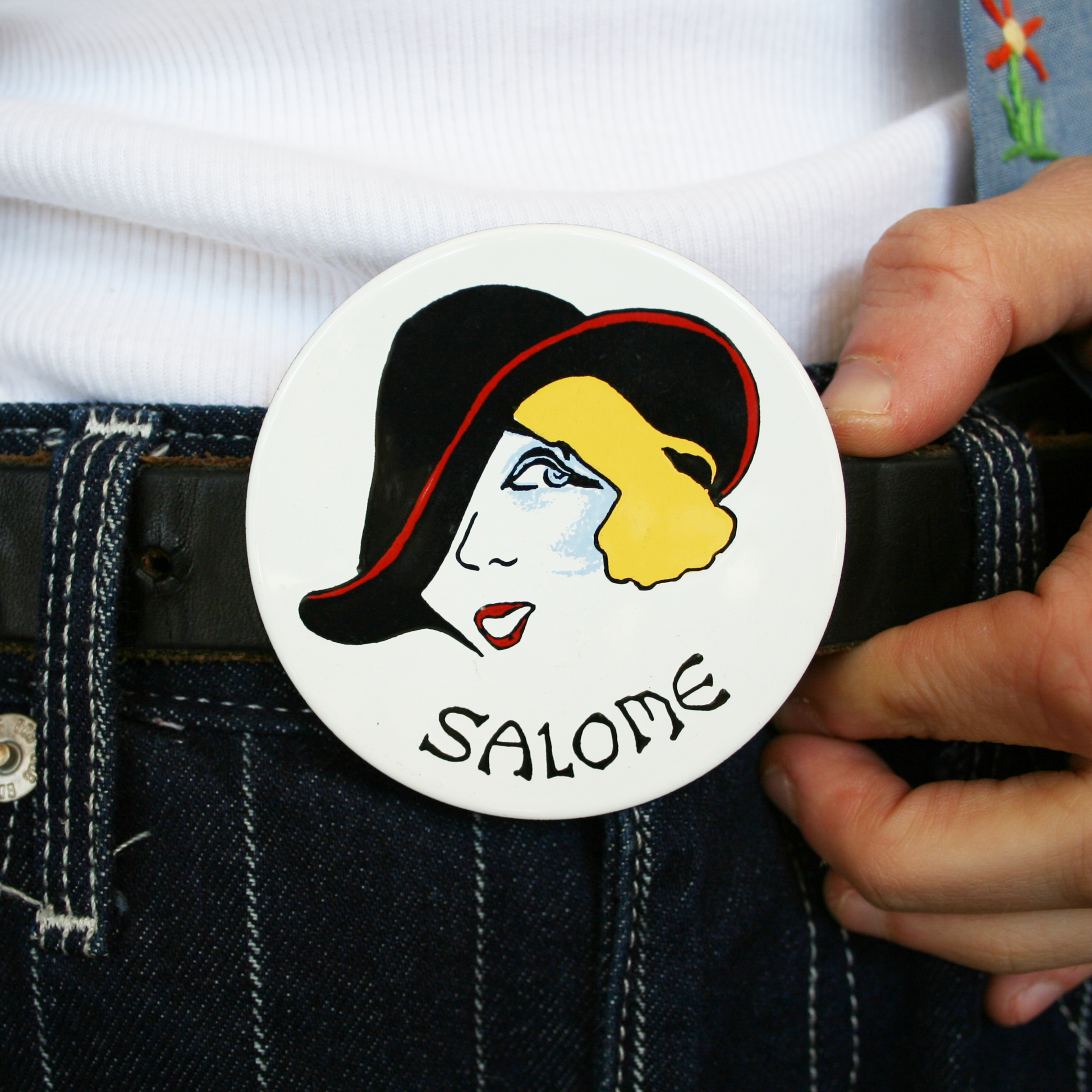 A hand holding a belt buckle with a vintage illustration of a woman named Salome wearing a black hat and white background