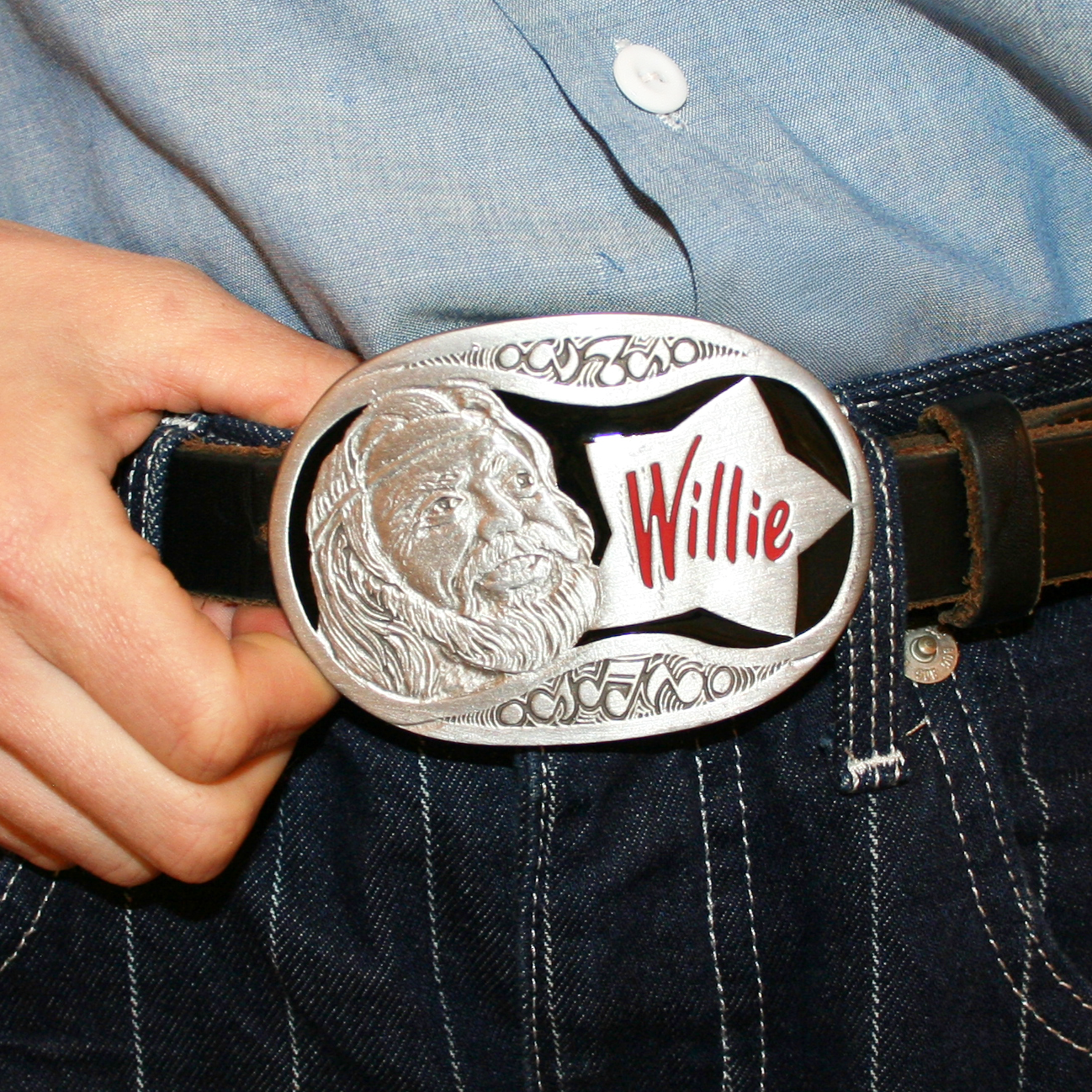 A person wearing a blue shirt and jeans with a vintage Willie Nelson belt buckle featuring a metal portrait of the artist.
