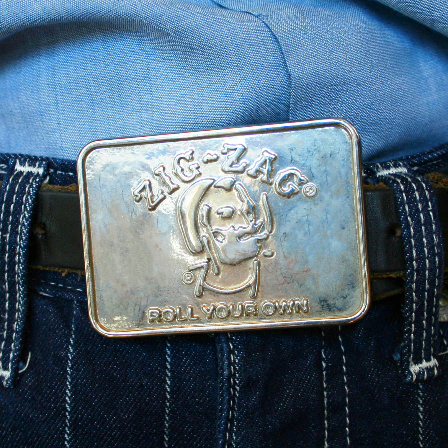 A vintage chrome-tone metal belt buckle with the Zig-Zag logo and the phrase 'Roll Your Own' embossed on it, attached to a blue belt and jeans.
