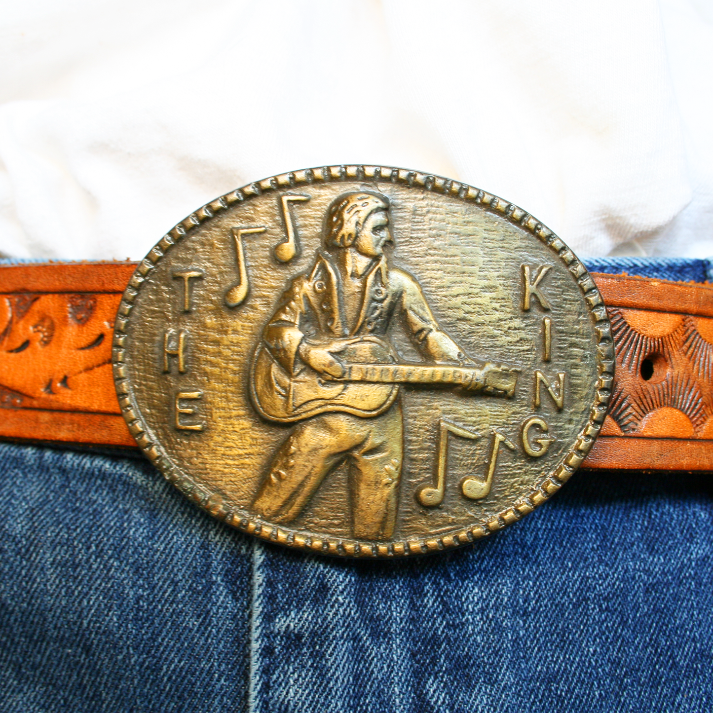 A brass belt buckle featuring an embossed design of Elvis Presley with a guitar, and the text 'THE KING' above.