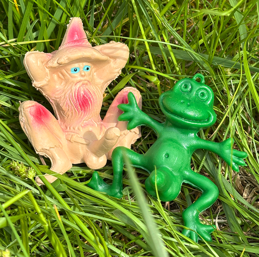 Two plastic novelty figurines, one pink and one green, designed to look like a well endowed toad and hillbilly character, placed in grass.