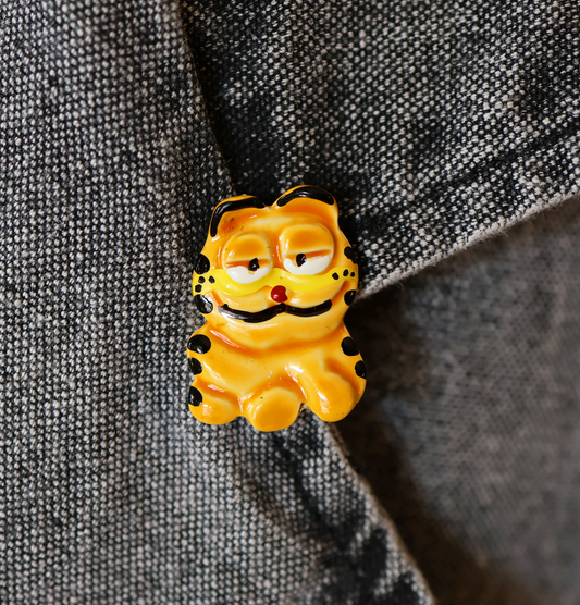 A yellow pin shaped like Garfield the cartoon character, with a whimsical expression, placed on a grey fabric surface.