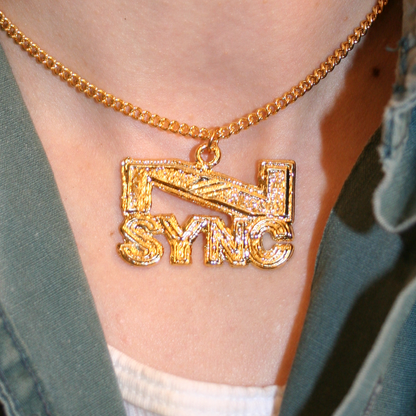 A close-up image of a vintage *NSYNC pendant necklace worn around the neck, featuring the band's name in bold gold-toned metal on a chain.