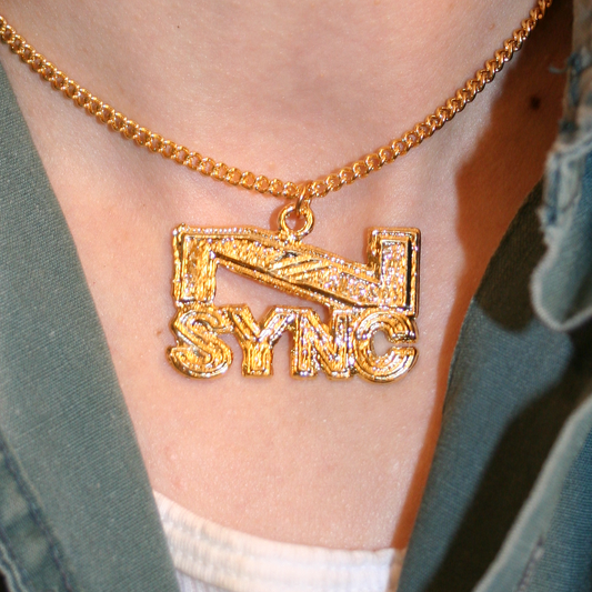 A close-up image of a vintage *NSYNC pendant necklace worn around the neck, featuring the band's name in bold gold-toned metal on a chain.