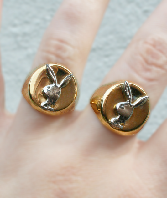 A close-up of two vintage Playboy Bunny rings with silver toned plating on a gold toned band, displayed on a person's finger.