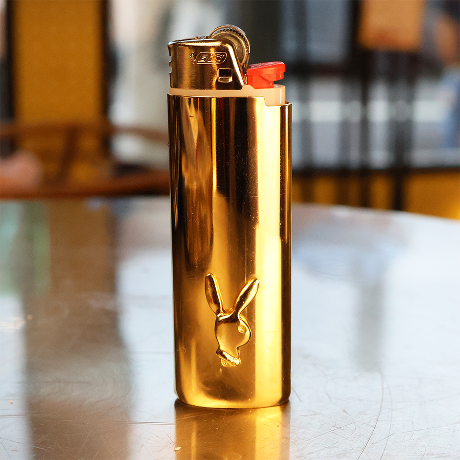 This image displays a stylish Deadstock Playboy Lighter Cover, characterized by its shiny, metallic gold finish. The cover features a prominent Playboy bunny logo engraved near the bottom.