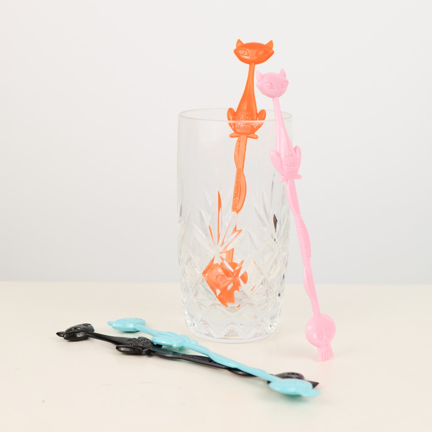 Clear glass with Shag, Josh Agle orange and pink cat-shaped drink stirrers on a light background
