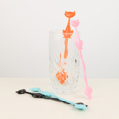 Clear glass with Shag, Josh Agle orange and pink cat-shaped drink stirrers on a light background