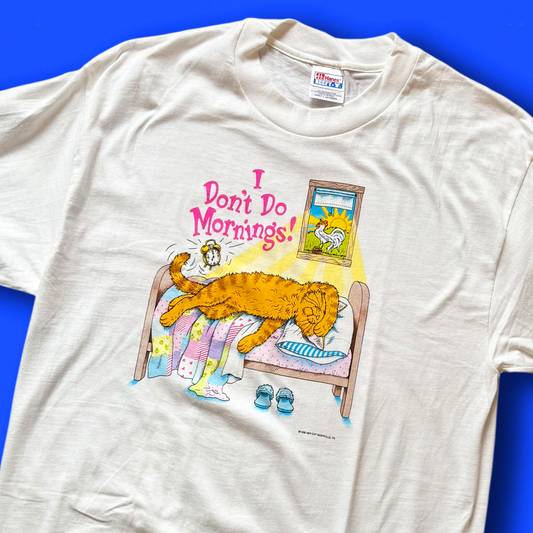 A vintage white t-shirt with a graphic print of a cat lying on a bed with the phrase 'I Don't Do Mornings!' and a depiction of a alarm clock and coffee cup. Hep cat
