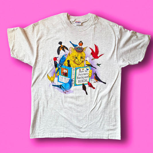 A vintage white t-shirt with a graphic print of a cat reading a book titled "how to attract backyard birds" surrounded by birds