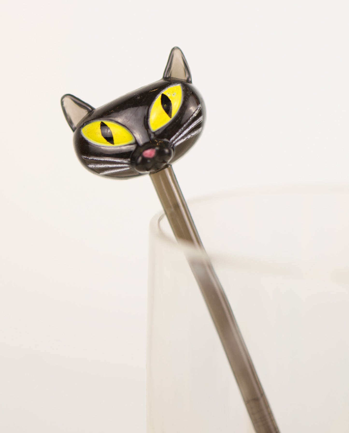Cat-shaped swizzle stick with yellow eyes and a pink nose on a white background. Vintage y2k Accoutrements 