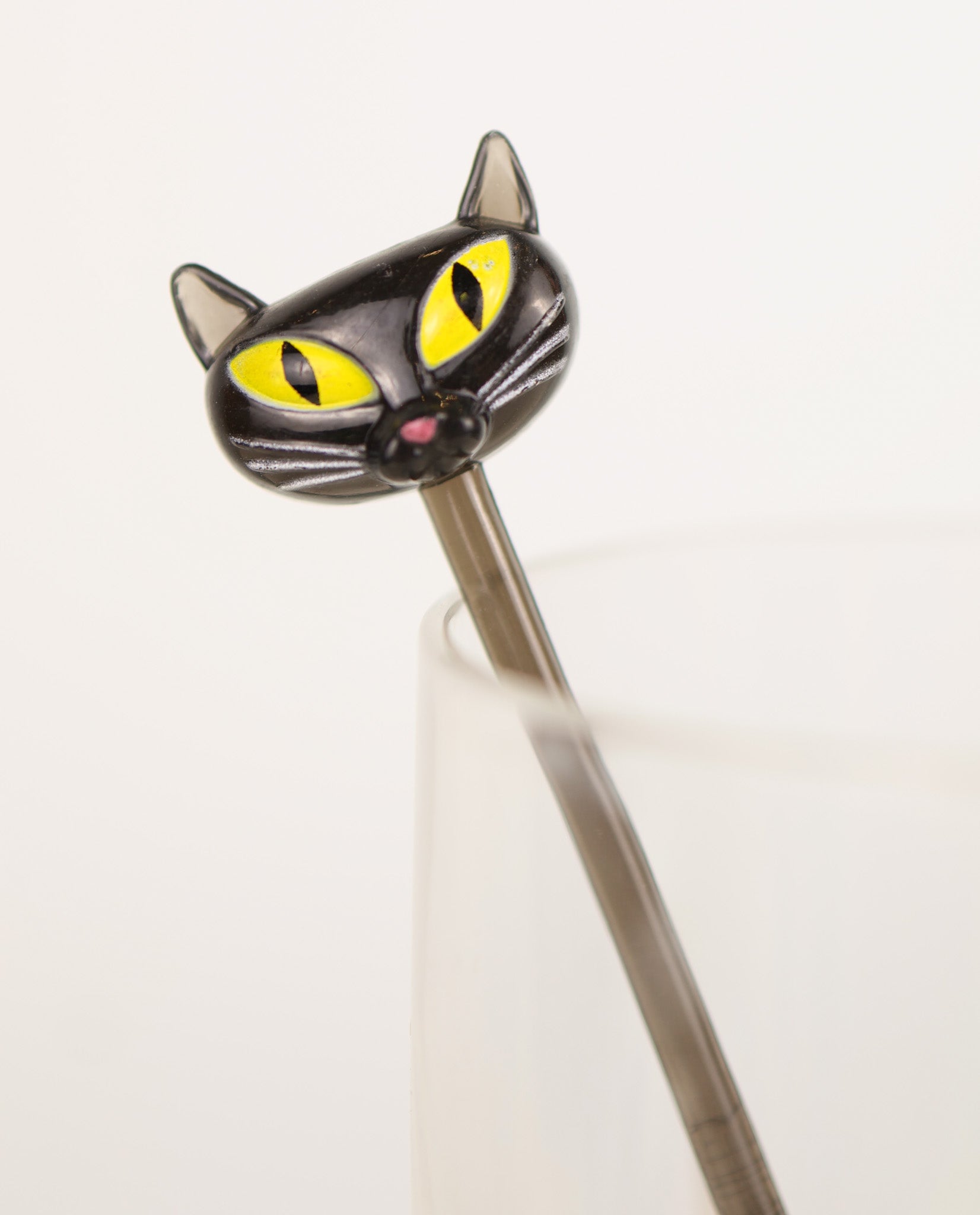 Cat-shaped swizzle stick with yellow eyes and a pink nose on a white background. Vintage y2k Accoutrements 